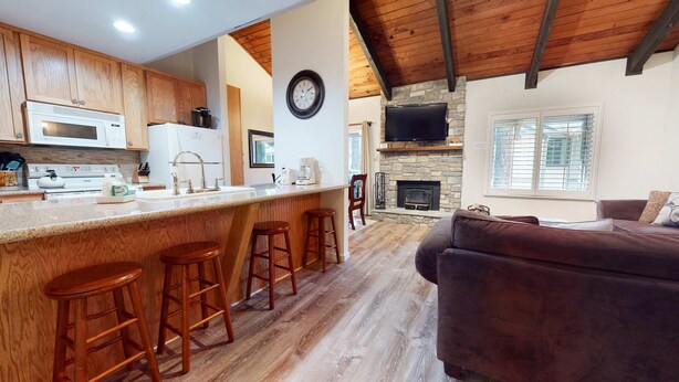 A kitchen with wooden cabinets, a microwave, and a sink. There are three wooden stools at the bar counter. A stone fireplace and a television are visible in the background.