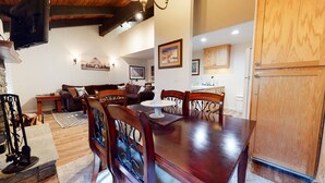 A dining area with a wooden table and chairs, a kitchen in the background, and a stone fireplace.