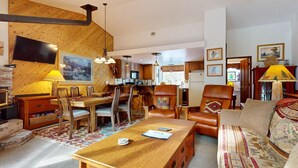 A cozy living room with a wooden dining table, leather armchairs, and a sofa. A kitchen is visible in the background with wooden cabinets and a window. - Mins to Lodge & Ski Shuttle | Parking for 2 (Mammoth Lakes)