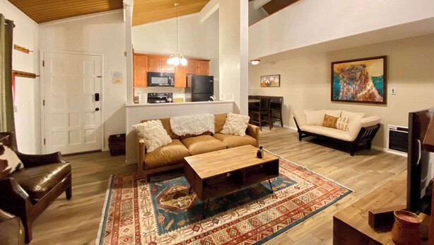 A spacious living area with a wooden ceiling, a kitchen in the background, and a large rug. - Sierra Manors #098 | Nearby Hike/Bike Trails (Mammoth Lakes)