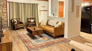 A living room with a brown leather sofa, two armchairs, a coffee table, and a patterned rug. A stone wall and wooden ceiling are visible. - Sierra Manors #098 | Nearby Hike/Bike Trails (Mammoth Lakes)