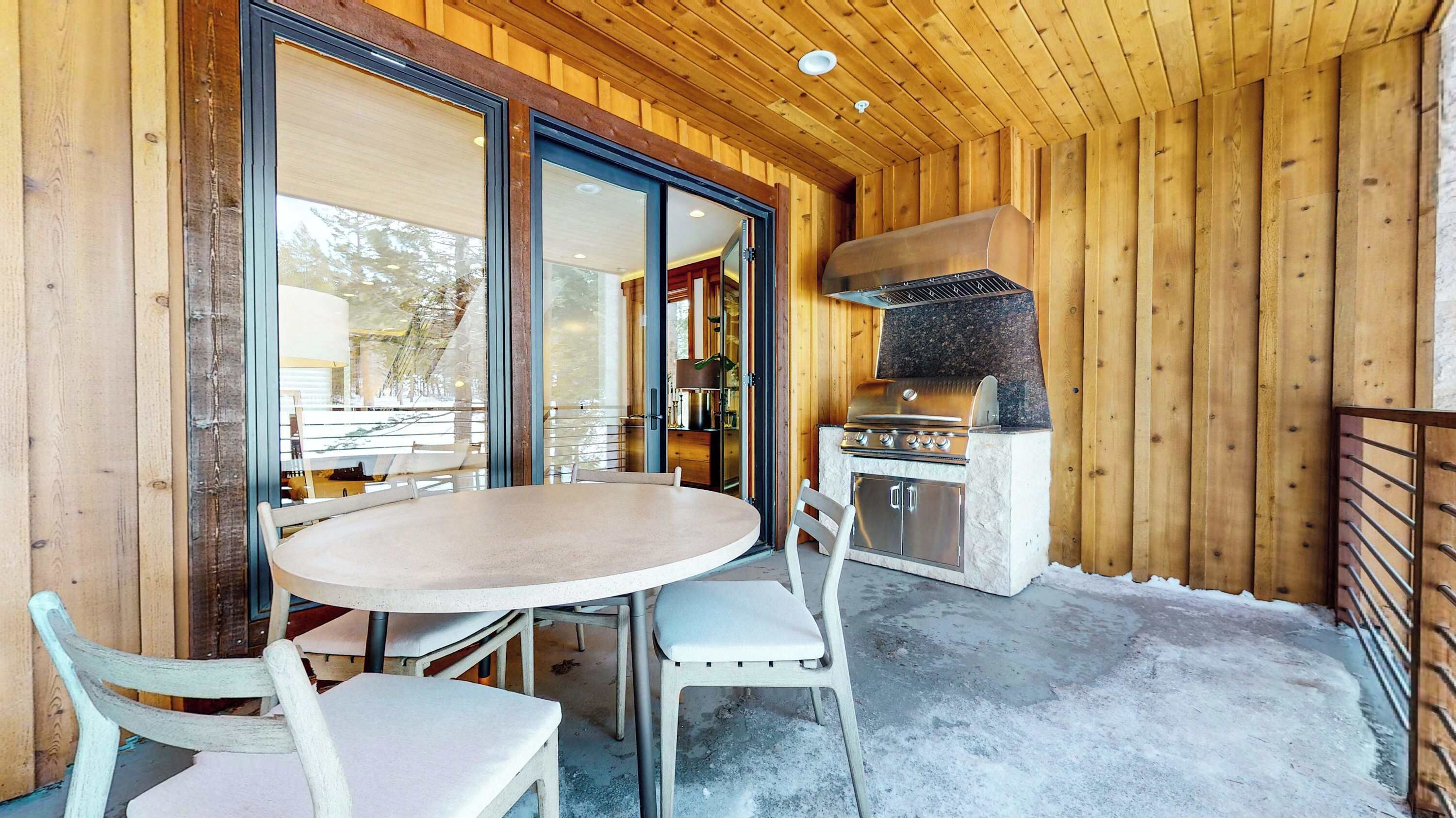 Una cabaña de madera con una mesa redonda y sillas, una cocina con una parrilla, y grandes puertas correderas de vidrio que dan a un espacio exterior.