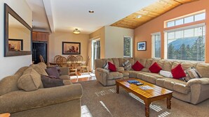 A spacious living room with a high ceiling, a dining area with wooden chairs, and large windows providing natural light.