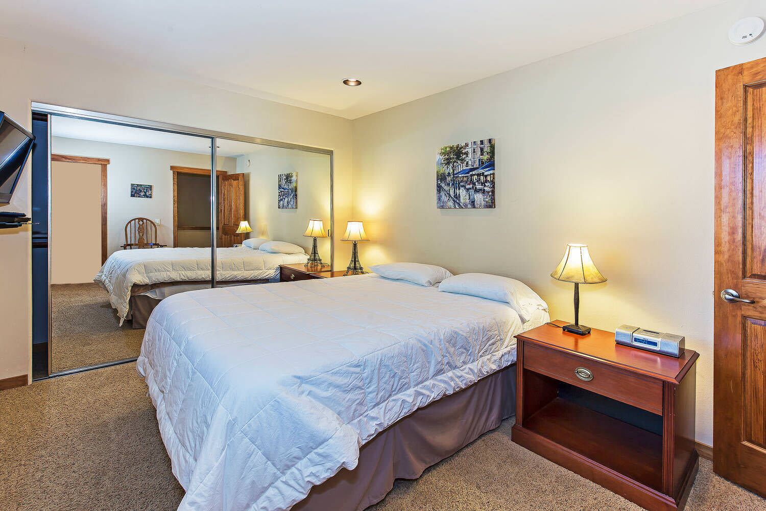 Een hotelkamer met twee bedden, een grote spiegel, een houten dressoir en een bureau met een lamp en een klok.