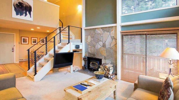 A spacious living room with a fireplace, a stone accent wall, a staircase, and a large window with blinds.