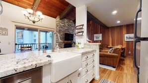 A kitchen with a stone fireplace, a sink, and a countertop. There is a wooden ceiling and a brown leather sofa in the living area.