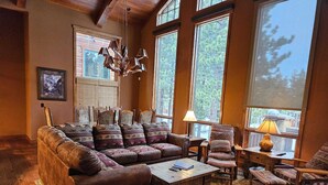 A cozy living room with a sectional sofa, a coffee table, and a patterned rug. There are large windows with a view of trees and a wooden ceiling. - Group Townhouse | 4 Ensuite Beds, Den, Sleeps 12 (Mammoth Lakes)