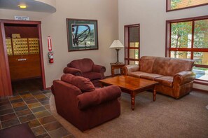 A lobby with a brown leather sofa, two armchairs, a wooden coffee table, a fire extinguisher, a framed picture, and a door with a sign. - Aspen Creek #204 | Near Golf, Fishing & Hiking (Mammoth Lakes)