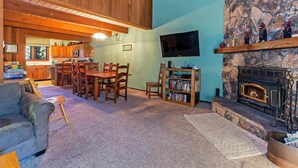 A cozy living room with a stone fireplace, wooden ceiling, and a dining area with chairs and a table.