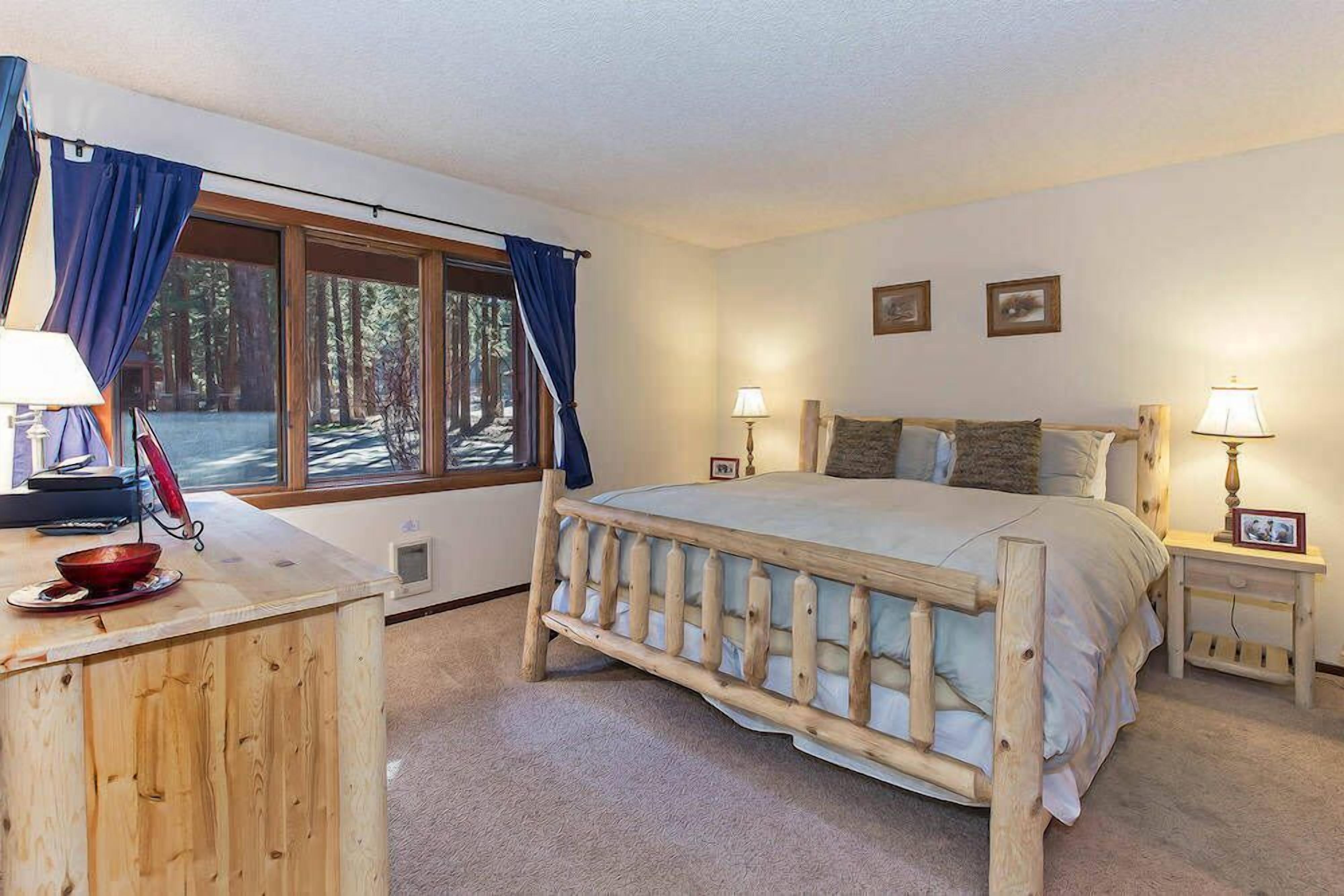 A bedroom with a wooden bed, a wooden desk, a window with curtains, and a nightstand with a lamp.