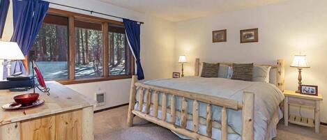 A bedroom with a wooden bed, a wooden desk, a window with curtains, and a nightstand with a lamp.