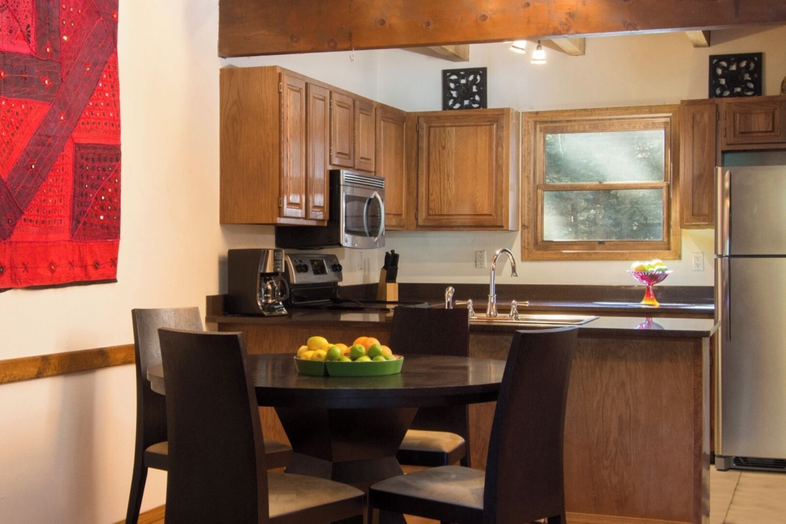 A kitchen with wooden cabinets, a stainless steel refrigerator, a microwave, and a coffee maker. There is a dining table with chairs and a bowl of fruit.