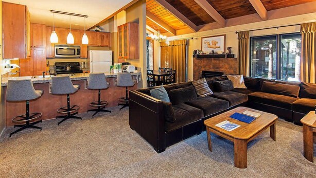 A cozy living area with a sofa, coffee table, and bar stools. A kitchen with a microwave and refrigerator is visible in the background.