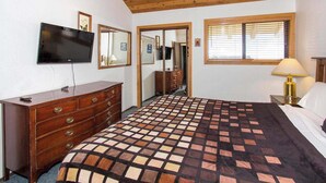 Ein Schlafzimmer mit Holzbalkendecke, einem an der Wand befestigten Fernseher, einem Schminktisch, einem Bett mit gemustertem Überwurf und einem Fenster mit Jalousien.
