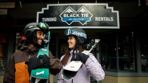Dos personas sosteniendo esquíes frente a un letrero de Black Tie Ski Rentals.