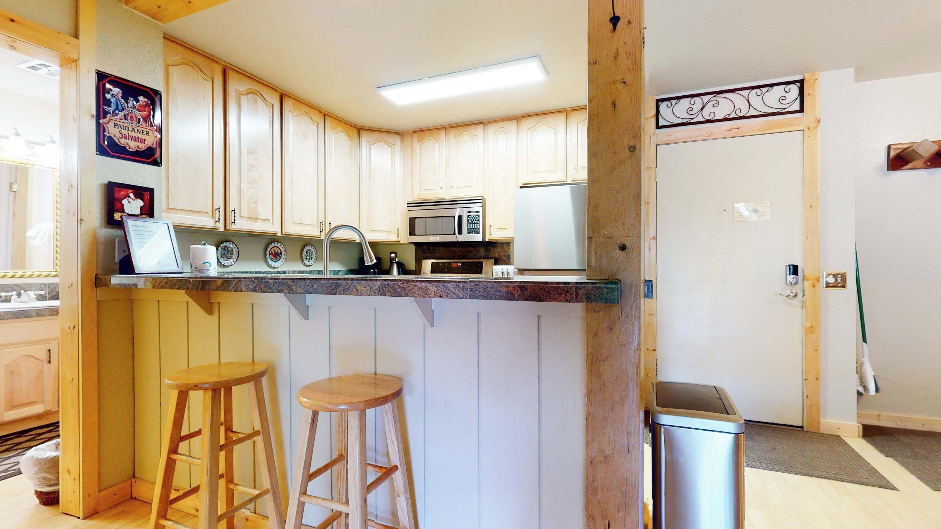A kitchen with wooden cabinets, a microwave, and a sink. There are two wooden stools and a trash bin.