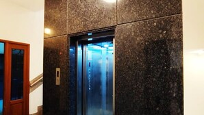 A modern elevator with a glass door, set in a hallway with a marble floor and a wooden door to the left. - Neighbour Inn Elite (Chennai)