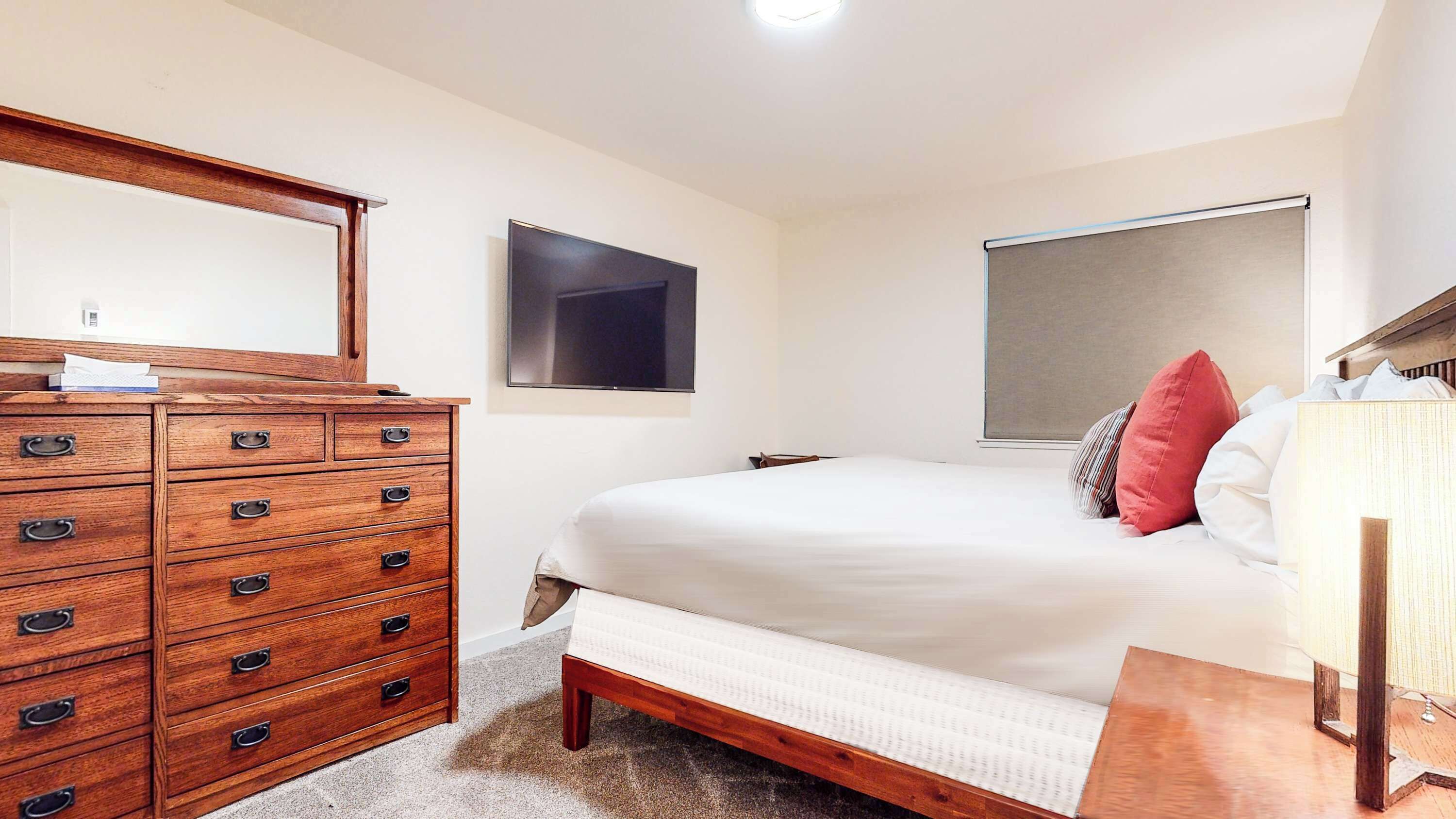 A bedroom with a wooden dresser, a bed with white bedding, a flat-screen TV, and a window with a blind.