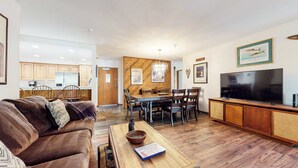 Una sala de estar moderna con un sofá, una mesa de centro y un televisor. Se observa un comedor con una mesa de madera y sillas. La cocina se ve al fondo, con armarios de madera.