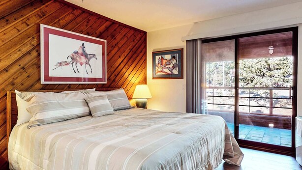 Un dormitorio con techo inclinado de madera, una cama con ropa de cama a rayas, dos obras de arte enmarcadas, una mesita de noche con lámpara y un ventanal corredero que da a un espacio exterior.