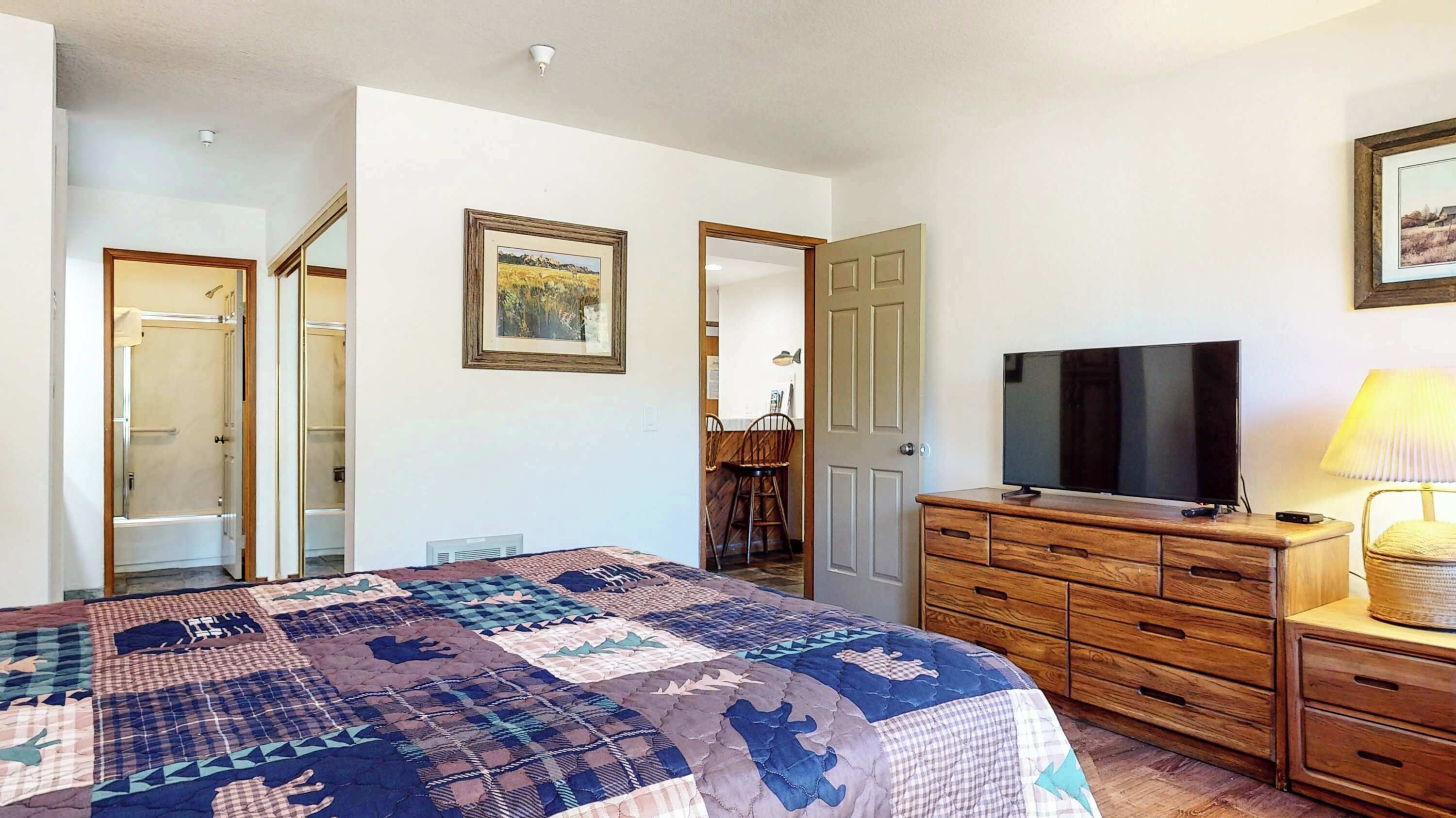A bedroom with a large bed, a dresser, a television, and a bathroom visible through an open door.