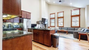 Una cocina amplia con encimeras de granito, armarios de madera y un refrigerador de acero inoxidable. Cuenta con una chimenea de piedra, una zona de comedor con sofá gris y ventanas grandes con persianas.