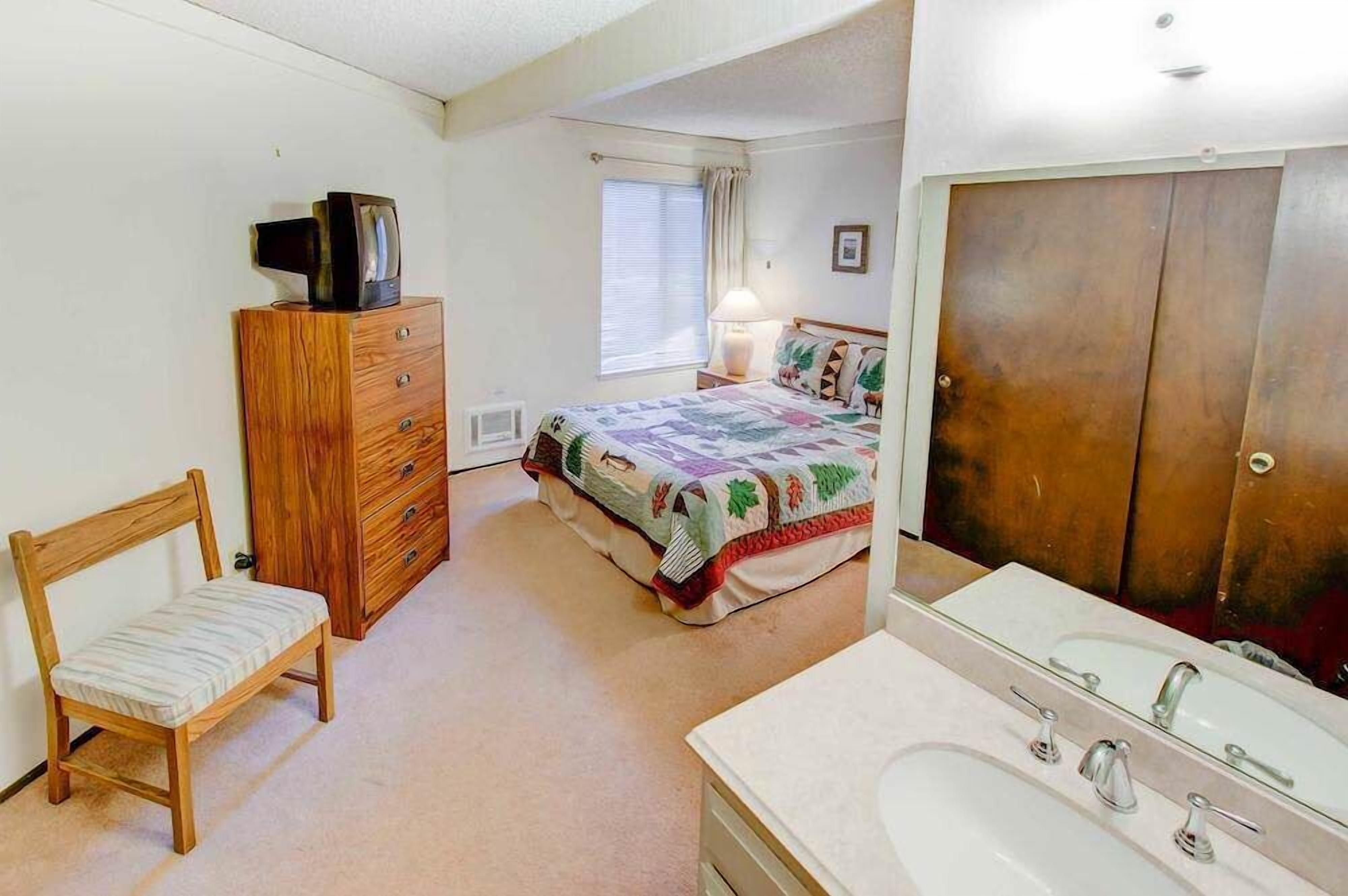 Un dormitorio con cama, cómoda, silla, televisor, ventana con cortinas y baño con lavamanos y espejo.