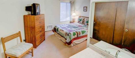 Un dormitorio con cama, cómoda, silla, televisor, ventana con cortinas y baño con lavamanos y espejo.