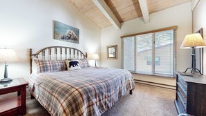 A bedroom with a bed, two bedside tables, a window with blinds, a mirror, and a painting on the wall.