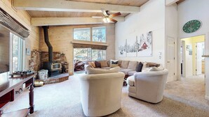 A cozy living room with a fireplace, a large window, a ceiling fan, and a clock on the wall.