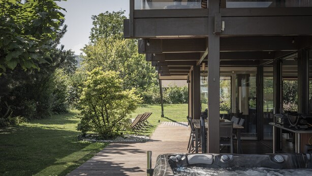 A hot tub in a covered outdoor area with a wooden deck, surrounded by greenery and a view of a house.