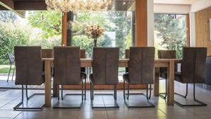 A modern dining area with a wooden table and six chairs, a chandelier, and a view of greenery outside.