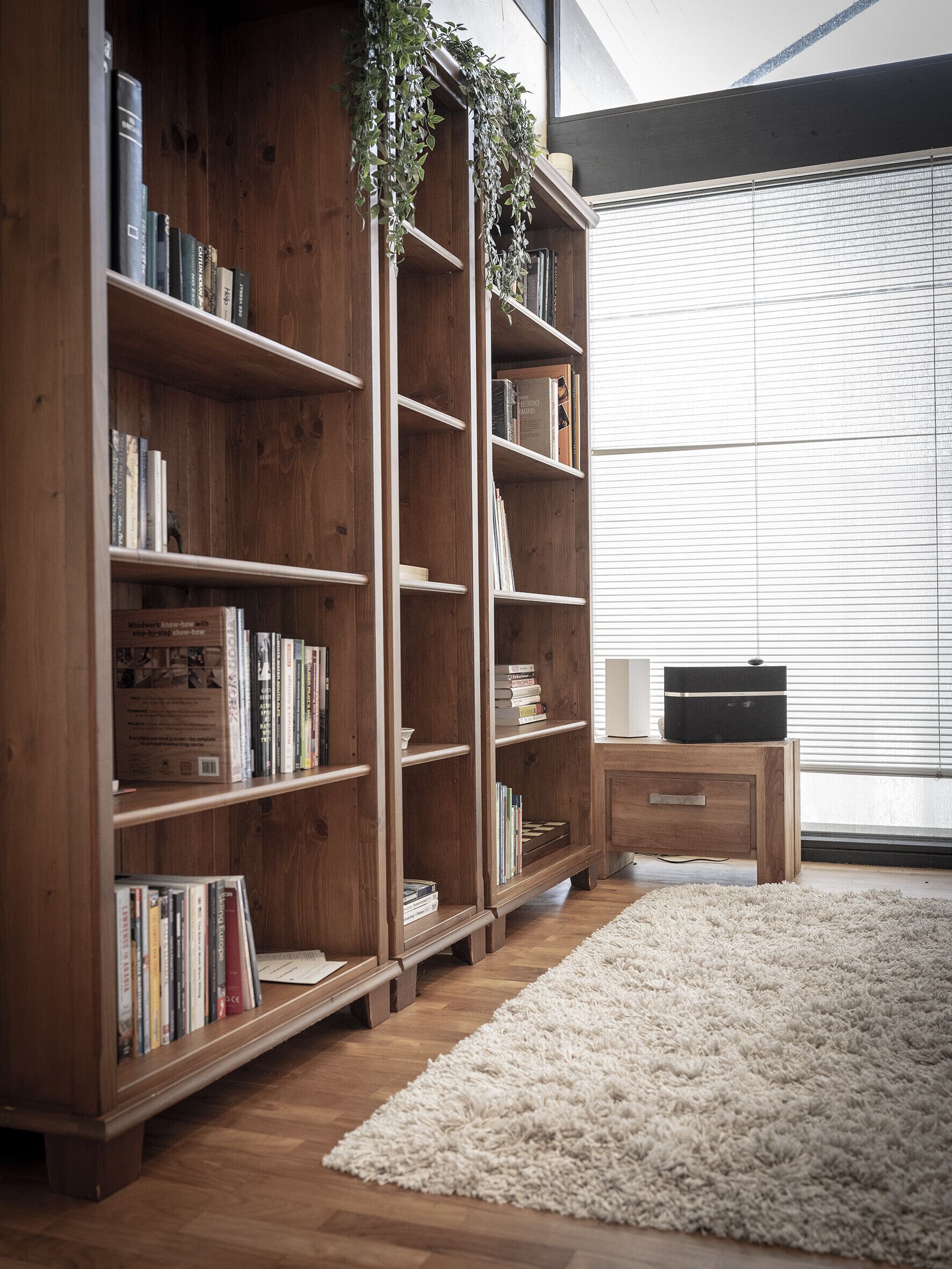 A wooden bookshelf filled with books, a white fluffy rug, and a small wooden table with a speaker.
