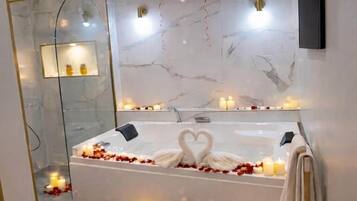 Une salle de bain moderne avec un mur en marbre, une baignoire décorée de pétales de rose et de bougies, et des ballons en forme de cœur.