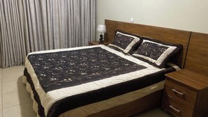 Een net opgemaakt bed met een bedspreidek en twee kussens, een houten nachtkastje met een lamp en een houten ladekast.