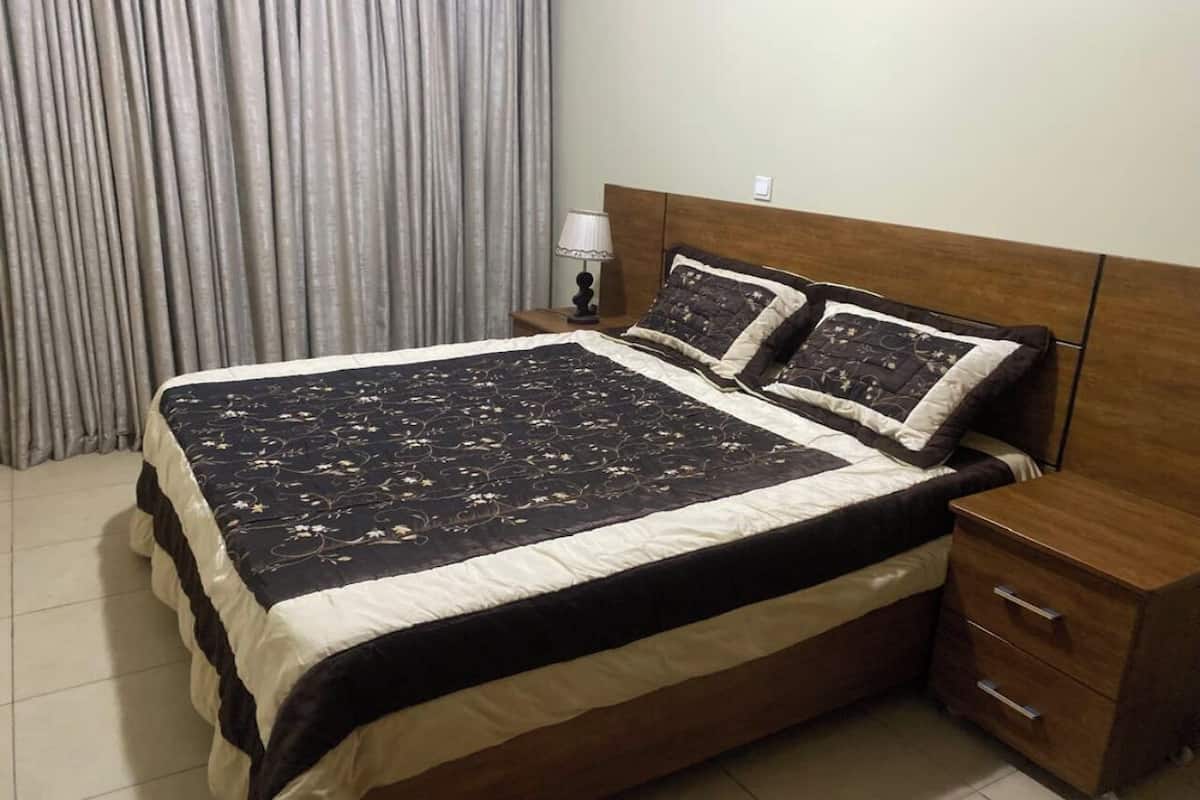 A neatly made bed with a patterned bedspread and two pillows, a wooden nightstand with a lamp, and a wooden drawer unit.