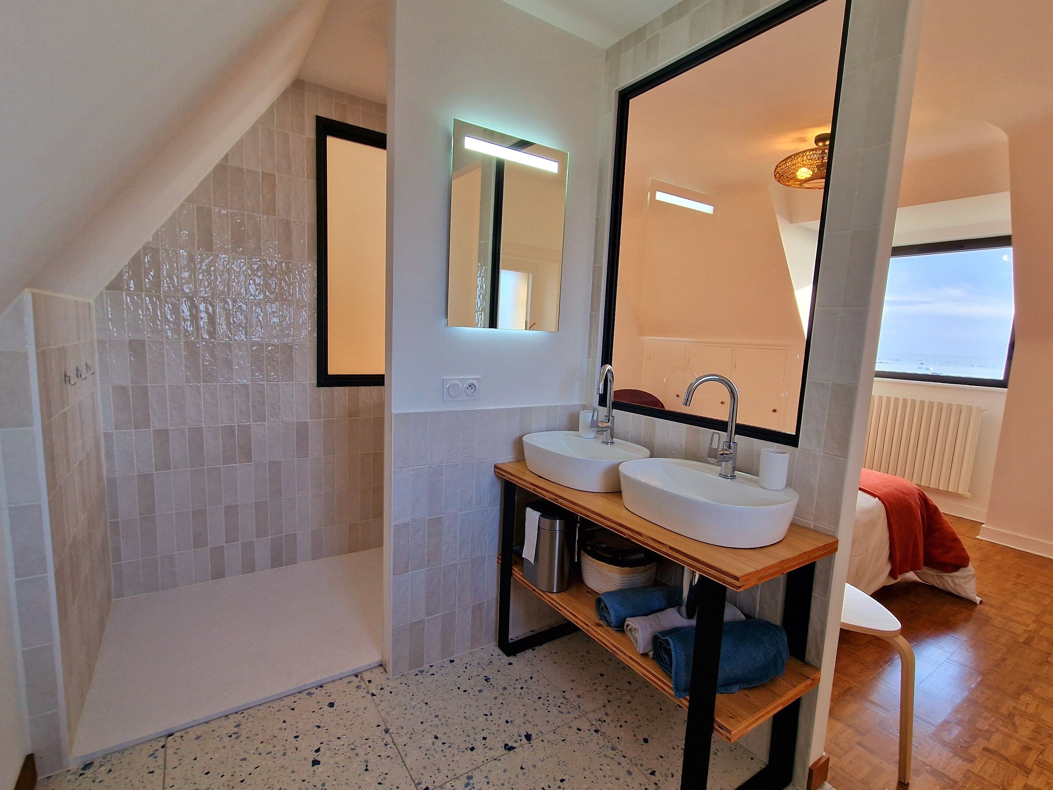 A modern bathroom with a shower, two sinks, a mirror, and a view of the sea.