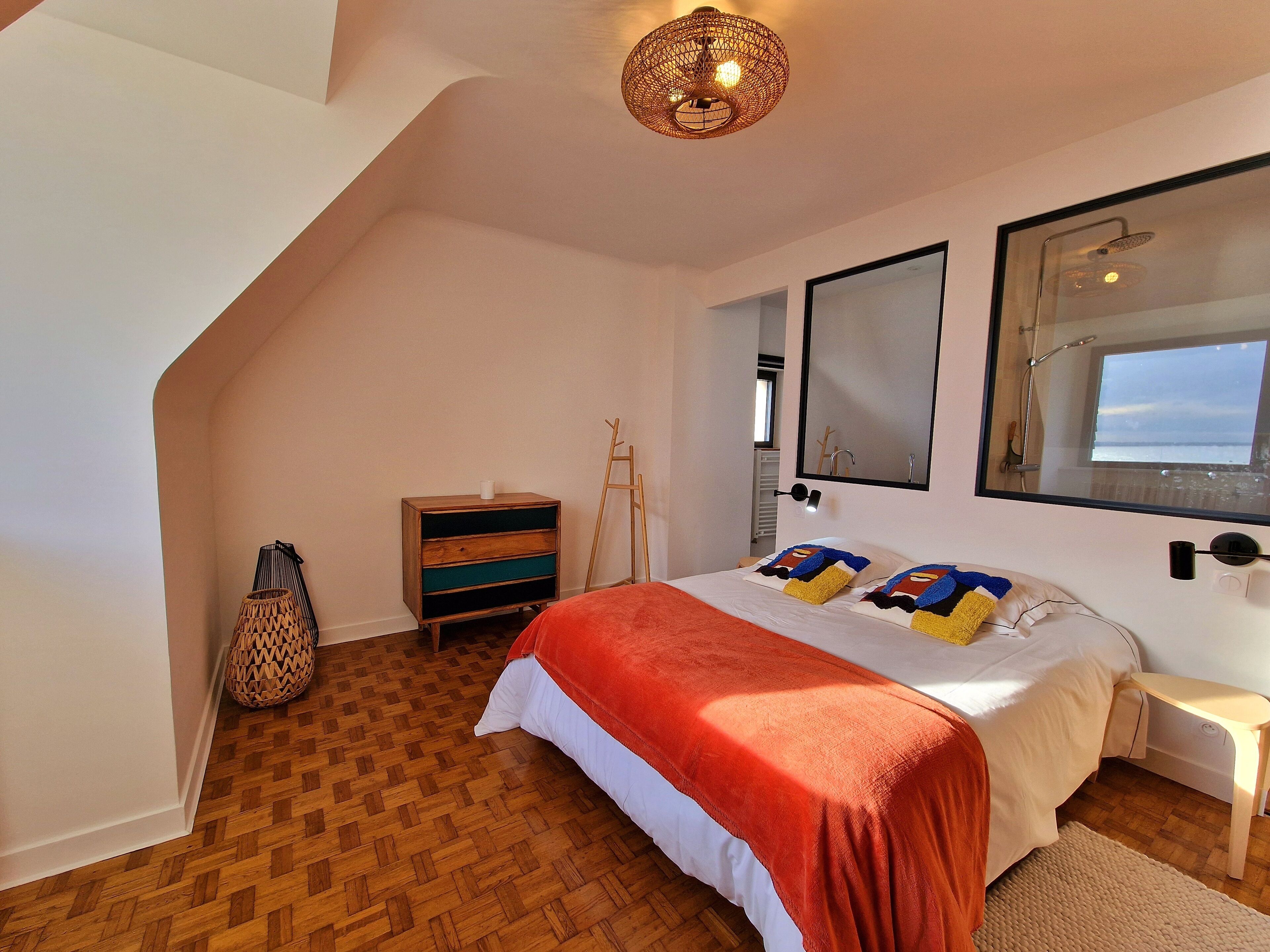 A bedroom with a sloped ceiling, a bed with a red blanket, a wooden dresser, a woven basket, and a large mirror reflecting a window view.