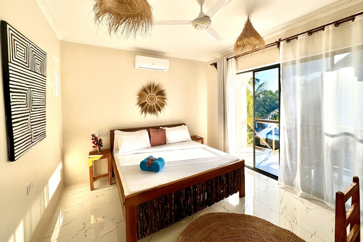 A bedroom with a bed, a ceiling fan, a decorative wall piece, a window with a view of a pool, and a woven chair.