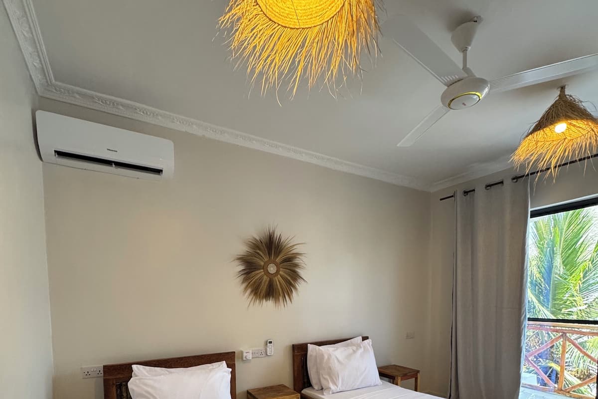 A hotel room with two wooden beds, a ceiling fan, a decorative wall piece, and a view of palm trees through the window.