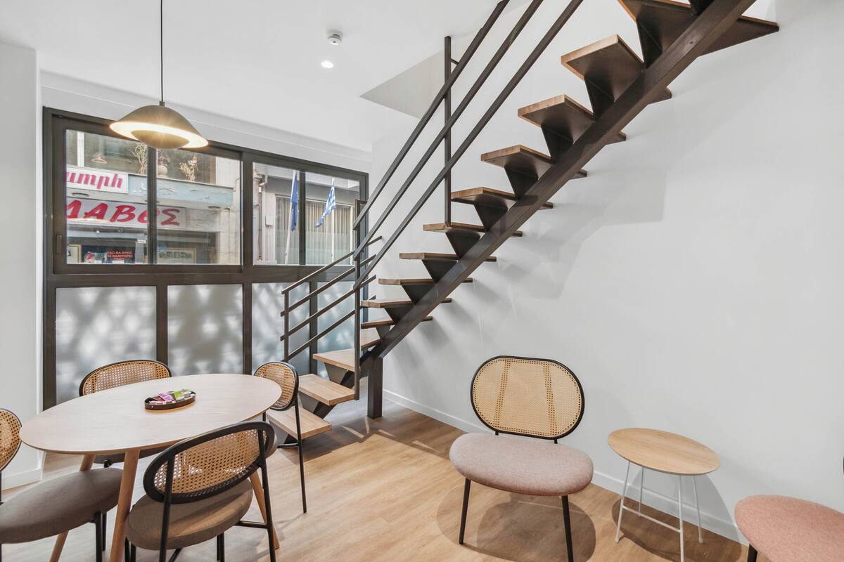 A modern interior with a wooden staircase, a dining area with wicker chairs, and a view of the outside through large windows.