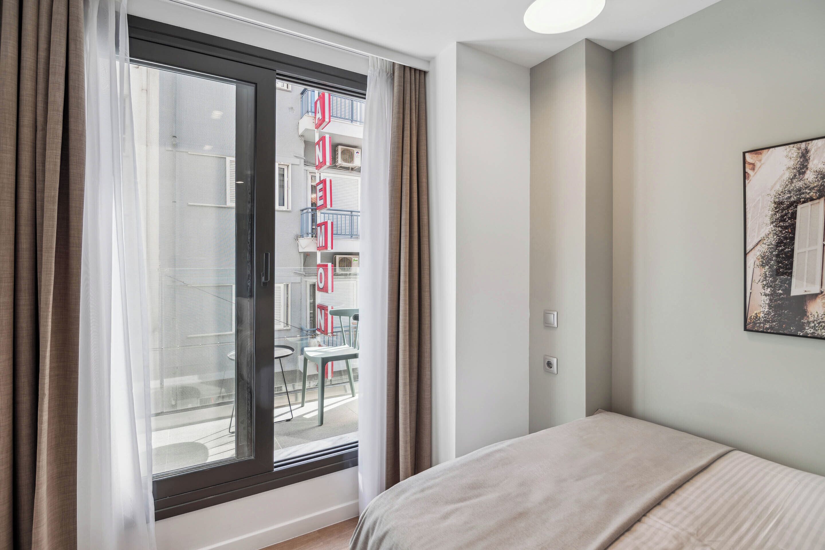 A bedroom with a large window, a bed, and a view of a balcony with outdoor furniture.