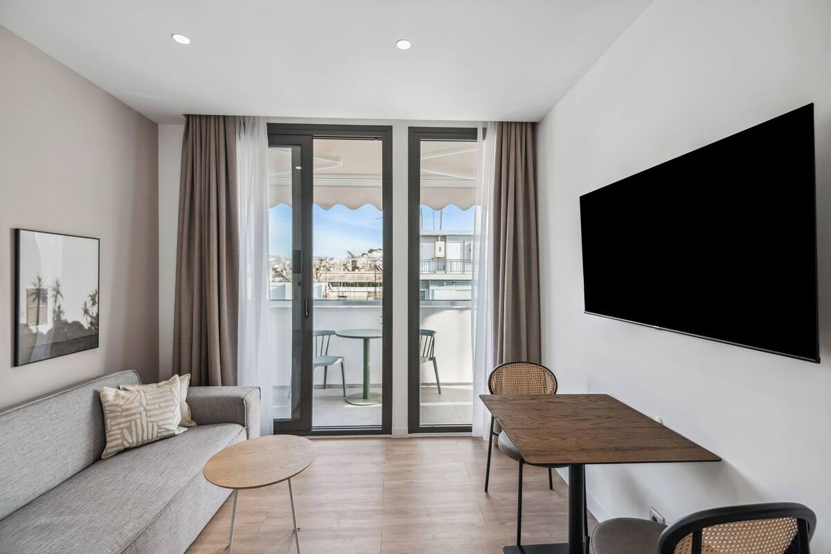 A modern living room with a sofa, a small round table, a dining table with chairs, a TV mounted on the wall, and a sliding glass door leading to an outdoor balcony.