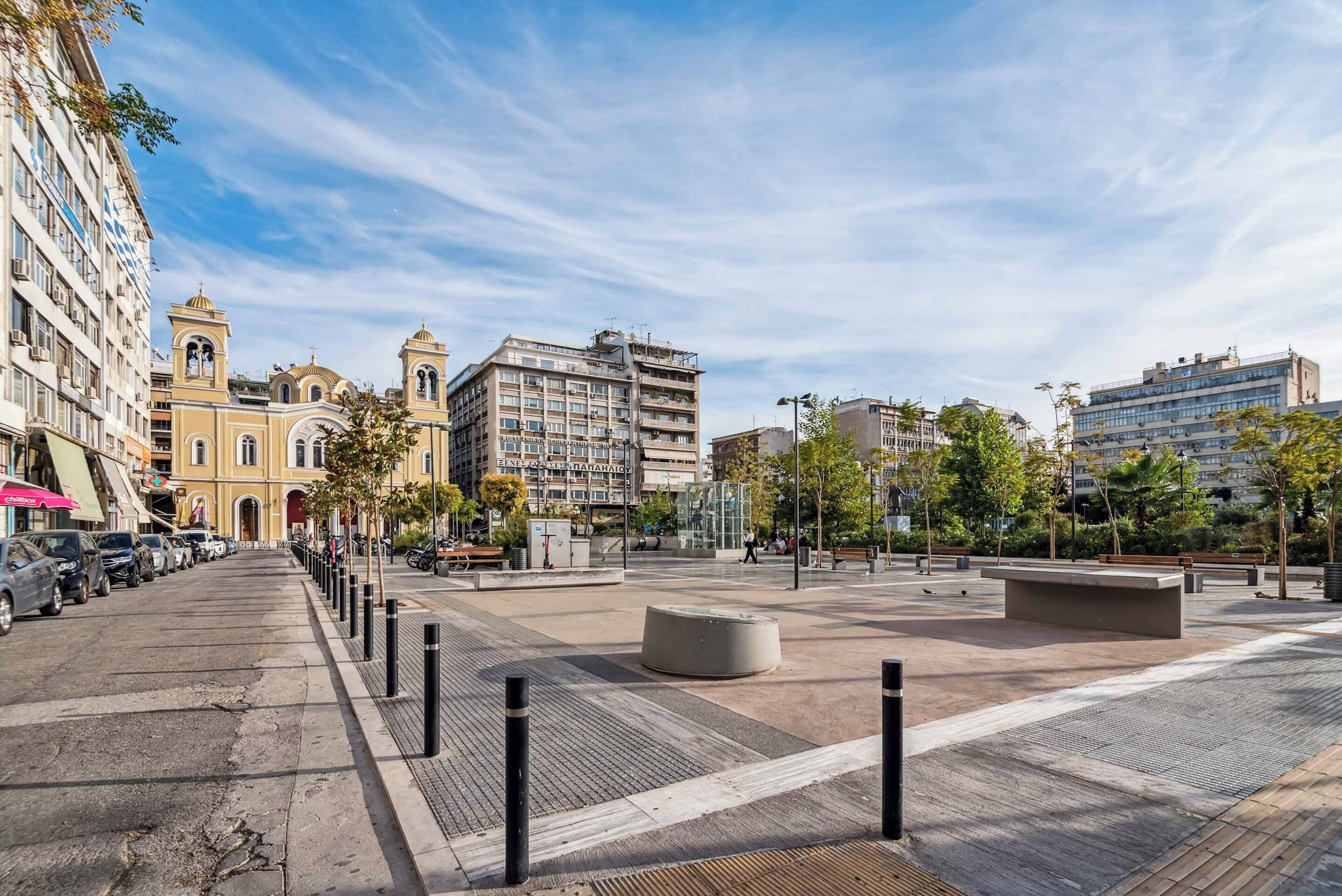 A spacious urban square with modern buildings, a prominent yellow church, and a clear sky.
