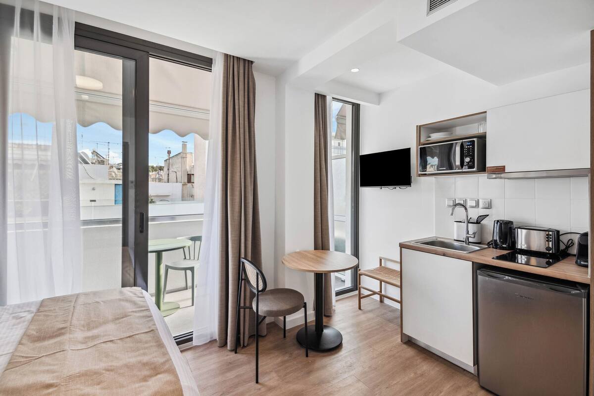 A modern hotel room with a bed, a small dining area, a kitchen with a microwave, and a TV.