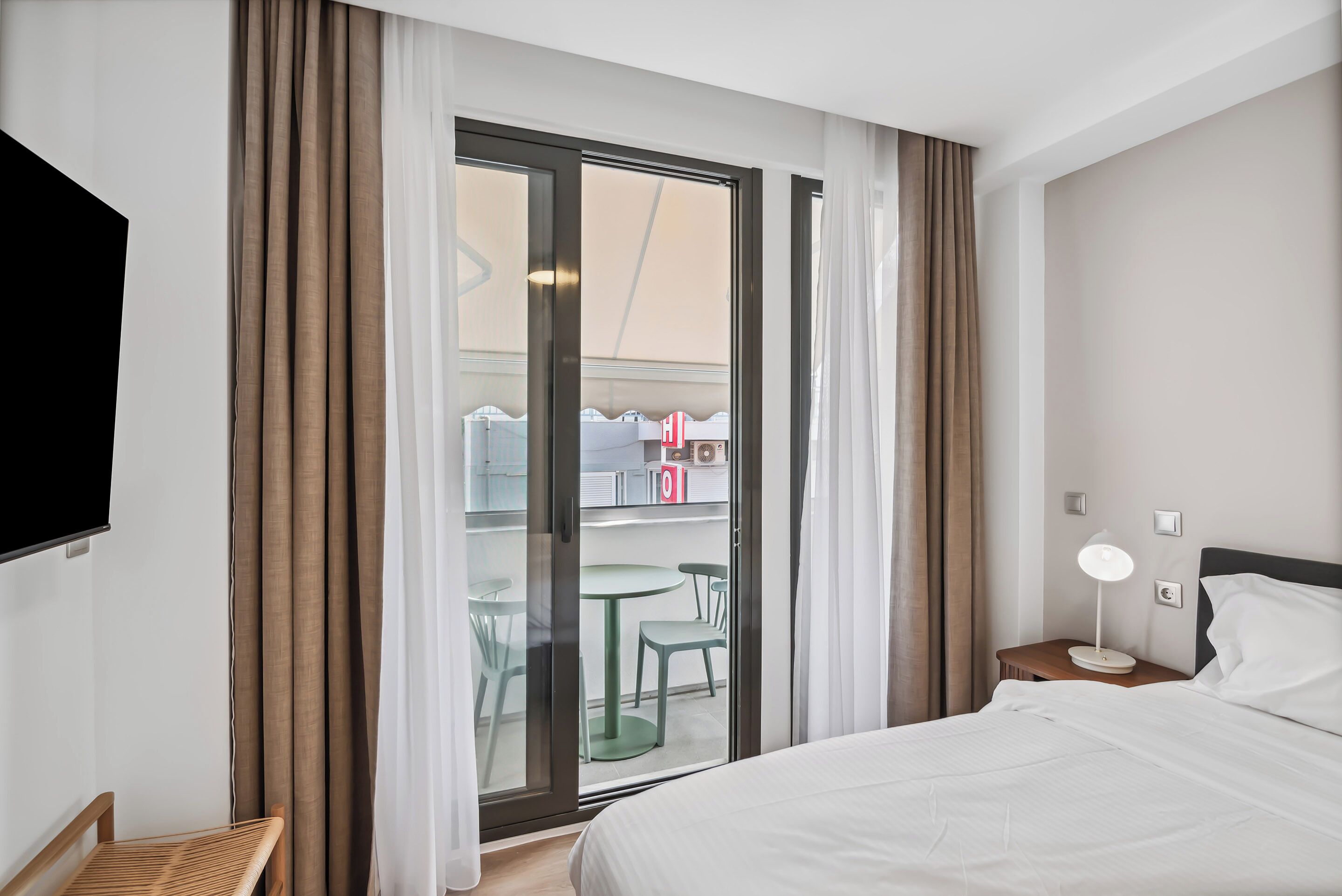 A hotel room with a bed, a nightstand with a lamp, a TV mounted on the wall, and a sliding glass door leading to a balcony with chairs and a table.