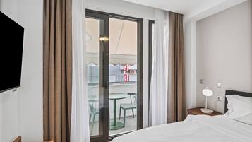 A hotel room with a bed, a nightstand with a lamp, a TV mounted on the wall, and a sliding glass door leading to a balcony with chairs and a table.