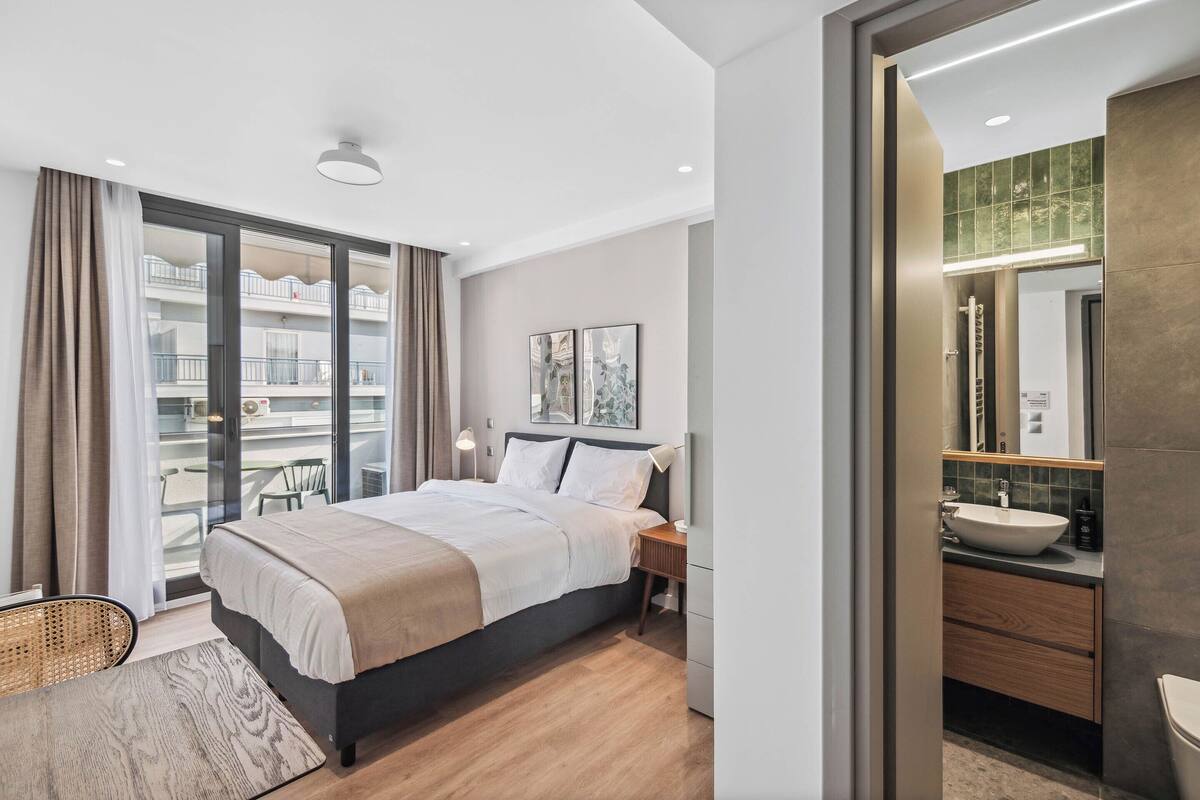A modern hotel room with a large bed, a bedside table, a bathroom with a sink and mirror, and a balcony with a view of the building.