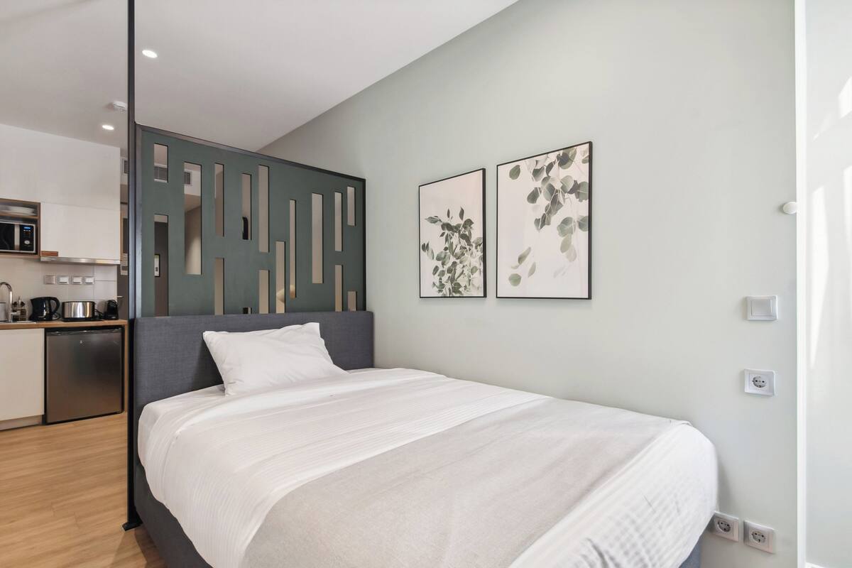 A modern bedroom with a bed, a kitchen area, and two framed artworks on the wall.