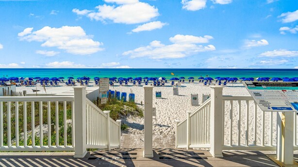 Une plage bordée d’une clôture en bois blanche, avec des parasols bleus et un ciel d’un bleu limpide.